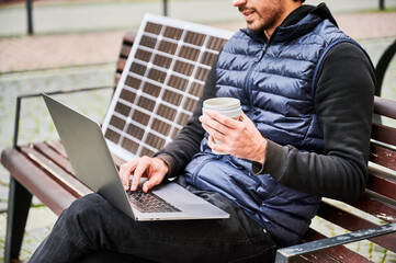 Cropped view of man sits on wooden bench in urban setting, working on laptop with cup of coffee in...