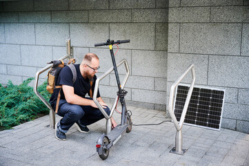 Man connects solar panel to electric scooter for charge, highlighting practical application of sustainable renewable energy. Concept of integration of eco-friendly technology in urban transportation.