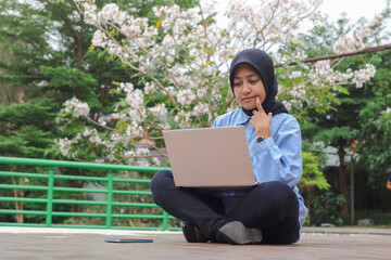 Portrait of Asian hijab woman sitting in city park, working on laptop