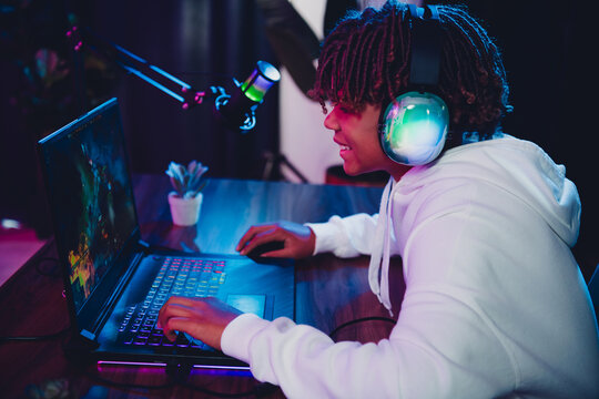 Young gamer live-streaming gameplay on laptop with illuminated headphones under colorful neon light in a casual home setup - Powered by Adobe