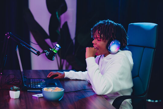 Young male gamer streaming gameplay and engaging with followers in a neon-lit setting using microphone and laptop