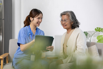 Asian Young female nurse in blue uniform smiling and showing digital tablet to elderly woman with...