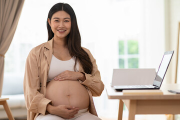 Asian Pregnant woman smiling and sitting indoors with hands on belly near laptop on table, natural...