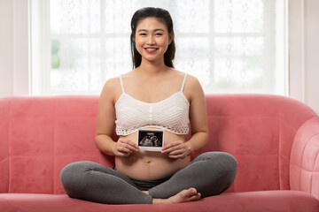 Asian Pregnant woman sitting cross legged on pink sofa holding ultrasound image with happy...