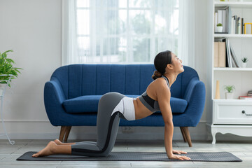 Asian Woman stretching on yoga mat in living room with blue sofa and white bookshelf