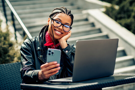 Lovely woman with braids smiling while using a smartphone and laptop outdoors on a sunny day in an urban environment