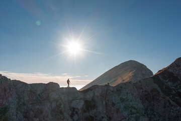 A lone hiker traverses a rugged mountain ridge as the sun shines brightly overhead, illuminating...