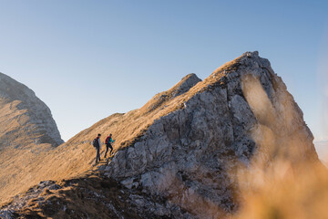 Two hikers traverse a rocky mountain ridge under a clear blue sky surrounded by nature.
