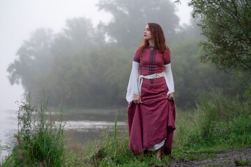 Ethereal woman in medieval dress gazes wistfully across a misty lakeside landscape.
