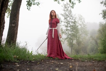 Mysterious woman in medieval dress stands pensively amidst a misty woodland path.