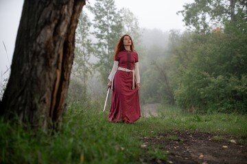 Mysterious woman stands amidst misty forest, clutching a sword.
