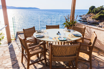 Scenic outdoor dining table on a sunlit terrace with panoramic sea views. Woven chairs and minimalist tableware create a relaxed yet refined atmosphere, ideal for vacation, hospitality.