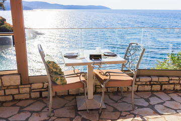 Intimate reserved dining table for two with elegant place settings, glass safety railing, and stunning direct ocean view at upscale Mediterranean waterfront restaurant terrace.
