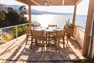 An inviting outdoor restaurant terrace with a wooden pergola and woven chairs, set against the backdrop of the sparkling blue Ionian Sea under the warm evening sun.