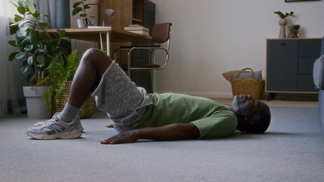 Full shot of mature African American man lying supine on floor carpet in living room, performing glute bridge exercise by lifting up hips and back and maintaining position