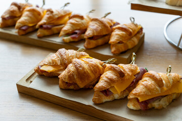 A wooden table displays neatly arranged croissants filled with ham and cheese, garnished with green picks. In the background, a tiered stand holds assorted sandwiches and pastries, creating a tempting