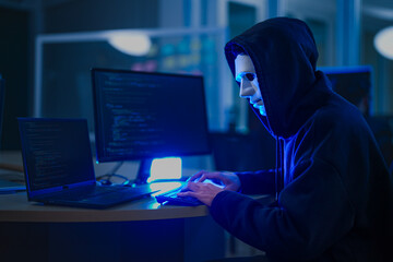 A mysterious hooded hacker working on a computer in a dark room, illuminated by blue screen light....