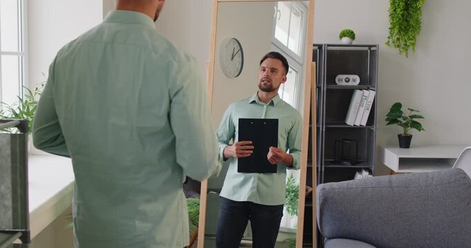 A man in a shirt holds a clipboard, studying his reflection during rehearsal and practice for a speech in a bright home office. He practices a speech before a mirror. Building confidence.