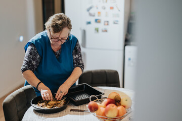 An elderly woman is preparing food using fresh ingredients in a cozy kitchen setting. A fruit bowl adds a touch of homely charm to the scene.