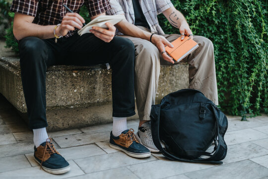 Two students sit on a bench outdoors reading books, emphasizing studying and education. A black backpack lies nearby on the pavement, surrounded by greenery, showcasing a relaxed academic environment.