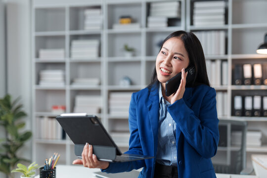 Smiling asian businesswoman talking on phone and holding tablet in office, she's wearing a blue suit and is standing in her office