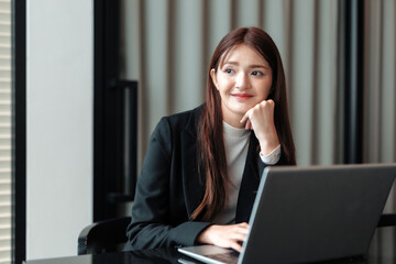 Young Asian businesswoman sitting at her desk, working intently on a laptop while contemplating...