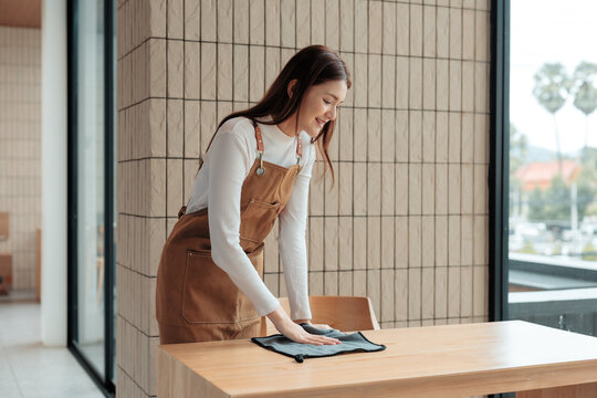 Young, smiling waitress cleaning a wooden table with a cloth in a modern cafe, creating a welcoming atmosphere for customers