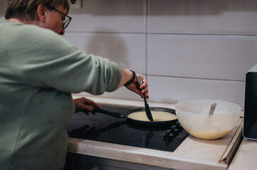 A senior woman wearing a green top cooking batter in a pan on an electric stove top, with a mixing...