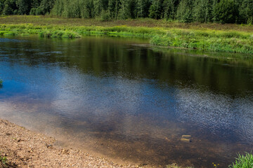 Peaceful riverbank with clear water and green forest reflection under blue sky. Natural summer landscape with calm ripples, grassy shore, and tranquil countryside scenery.