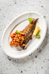 Grilled fish fillet with golden skin, served with chunky vegetable salsa, lemon wedge, and parsley on a white plate over a stone surface
