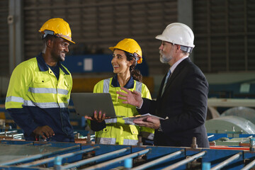 The workers are standing in a factory, looking at a tablet. The engineer talks with the woman's workers. They wear a yellow and white safety helmet for safety.