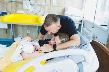A man hugs his wife and newborn baby in the delivery room. The woman gave birth and the man is...