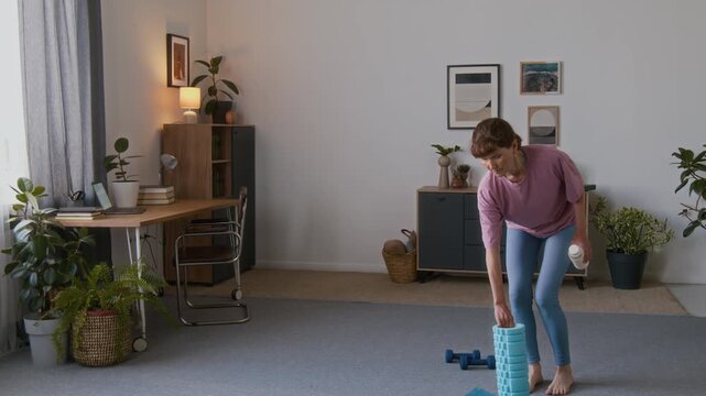 Wide lockdown shot of slim senior Caucasian woman in pink t-shirt, blue leggings walking into living room, carrying exercise gear and water bottle, standing on mat, warming up before exercise routine