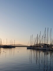 Sailboats docked in a marina capture the last light of sunset, their reflections shimmering on the calm water