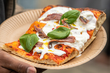 A slice of pizza topped with fresh basil leaves, anchovies, and melting stracciatella. The pizza is served on a paper plate, with the slice being held by a person