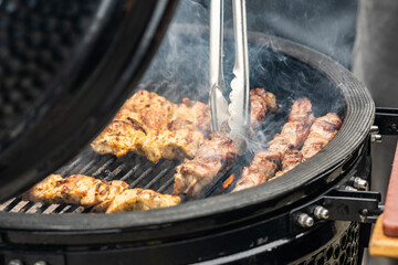A close-up shot of a person grilling skewers of meat on a barbecue, using grill tongs to flip the meat as smoke rises from the grill