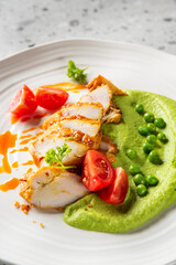 Grilled chicken breast with orange sauce, green pea puree, cherry tomatoes, and parsley on a white plate. Elegant gourmet presentation on a stone surface