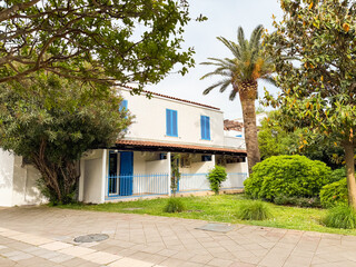 White building with blue shutters surrounded by green grass, palms and bushes. Architecture, urban housing, residential lifestyle and natural summer environment with balance.