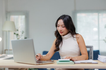 Asian businesswoman working with laptop computer, holding pen and looking at screen, sitting at desk in modern office with documents and notebooks