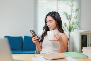 Smiling businesswoman drinking coffee while using smartphone in bright modern office, working remotely or enjoying break