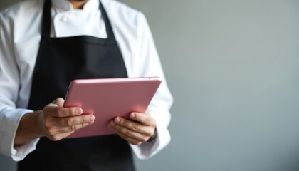 Chef in white uniform, black apron holds pink tablet, using digital device for cooking, restaurant management, kitchen innovation. Modern technology in hospitality industry for efficient service,