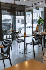 Empty modern cafe interior with wooden tables, black slatted chairs, large front windows, and view of parked cars outside on a cloudy day