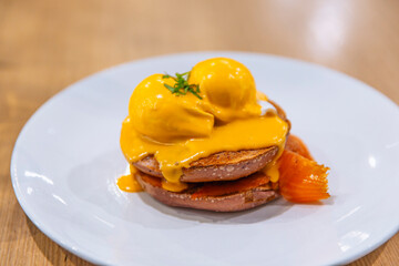 Stylish brunch dish of eggs benedict on purple bagel with smoked salmon and hollandaise sauce, served on a white plate on wooden table