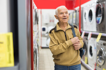 elderly grayhaired man pensioner looking washing machine at counter in showroom of electrical...