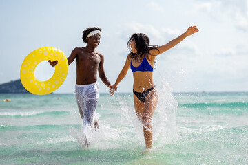 Happy interracial couple enjoying a fun beach day, splashing in the ocean waves, holding hands