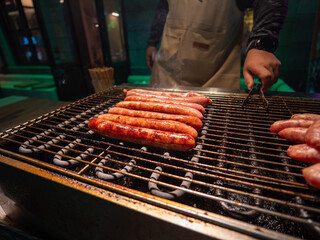 A person using tongs to grill Taiwanese sausages on a charcoal grill at a night market.