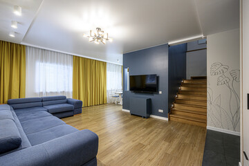 well-lit living room featuring a blue sectional sofa, wooden floors, yellow and white curtains, a TV mounted on a blue wall, and a wooden staircase
