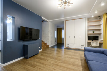 Modern living room with a blue accent wall, mounted TV, wooden floor, and a glimpse of a staircase. Minimalist design with white wardrobe and yellow curtains