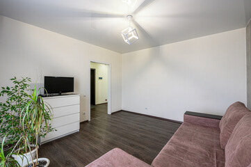 bright living room features a brown sofa, white dresser with a TV, and indoor plants, with a doorway leading to another room