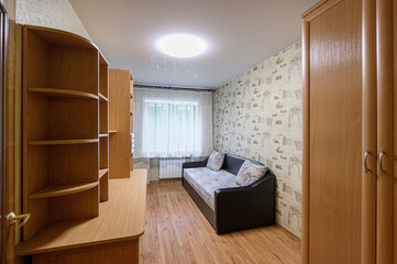 cozy room features a desk with shelves, a sofa with cushions, and a window with sheer curtains. Neutral wallpaper and wooden floors complete the scene